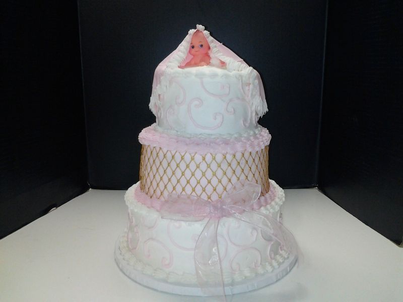 Three-tiered baby shower cake; white frosting, pink accents, decorative lattice, and baby figurine on top.