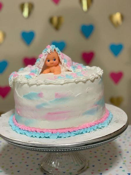 Gender reveal cake with baby figurine, decorated in pink and blue frosting, on a cake stand.