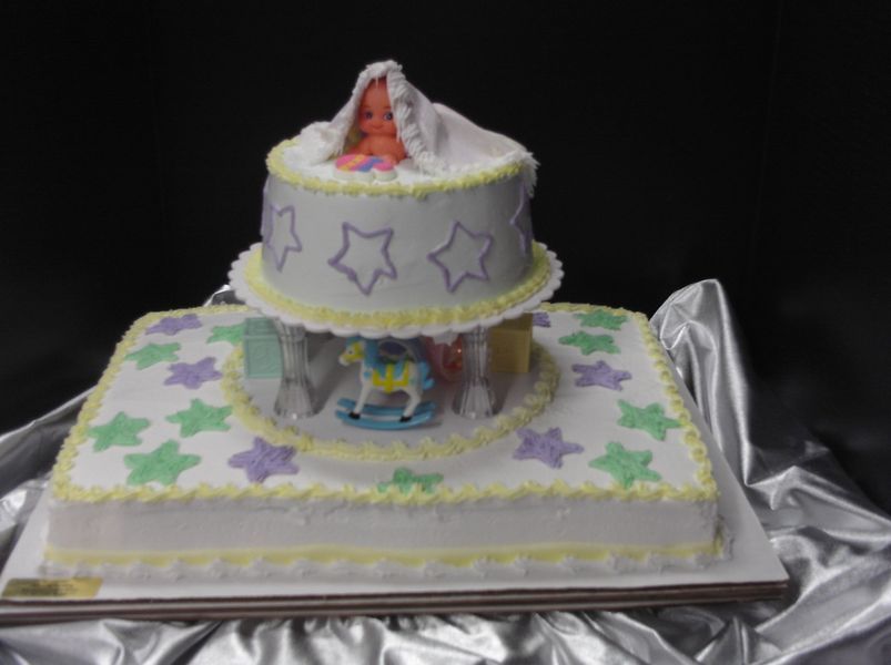 Two-tiered baby shower cake with toy baby, stars, rocking horse, and pastel frosting.
