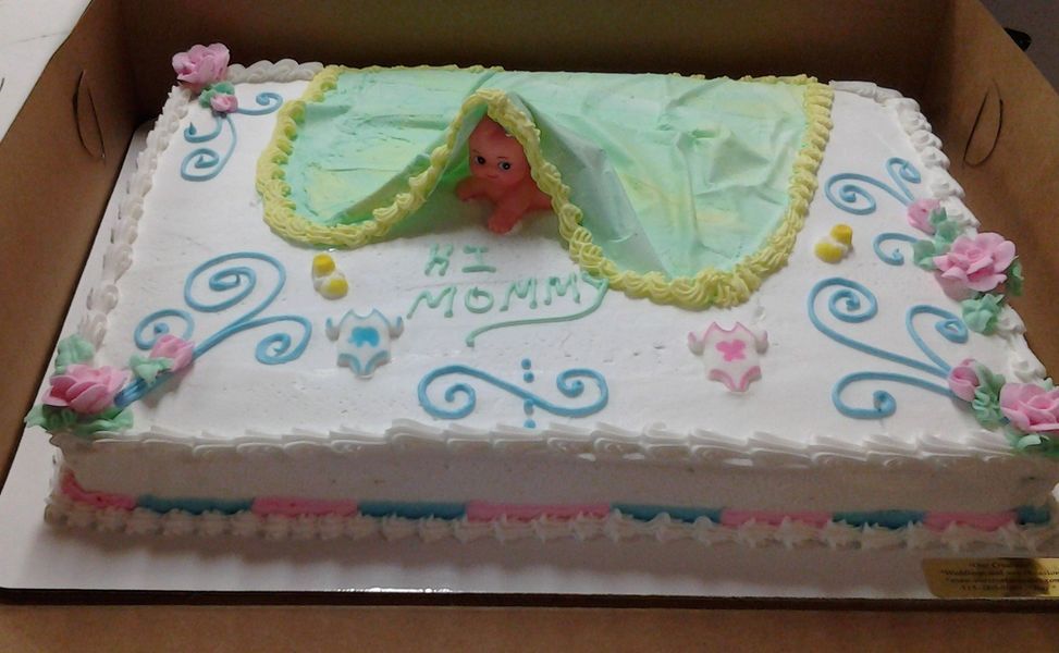 Cake decorated for a baby shower. White frosting, baby under a green blanket, pink and blue accents, text 