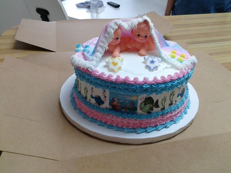 Baby shower cake with pink and blue frosting, featuring baby figurines under a blanket.