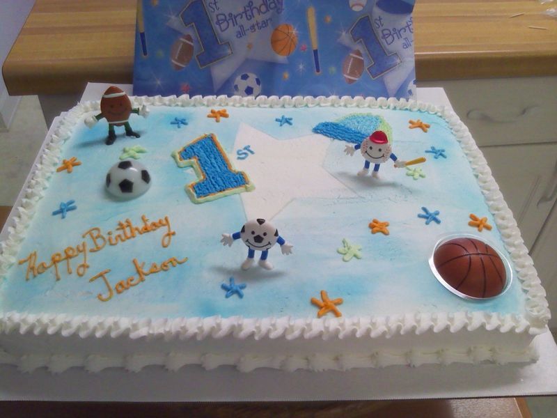 Rectangular cake with blue frosting and sports-themed decorations for a first birthday. 