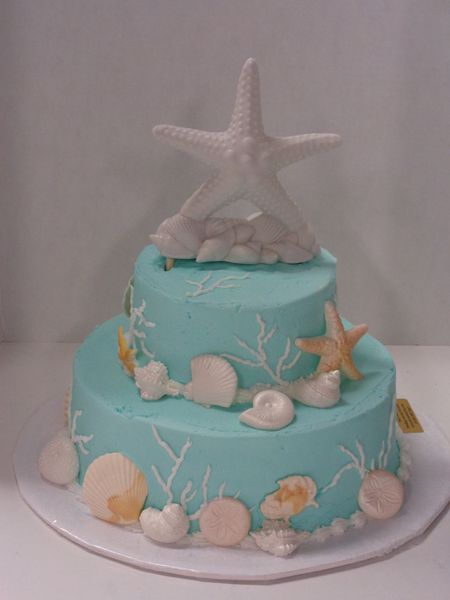 Two-tiered cake with blue frosting, decorated with seashells, starfish, and white coral.