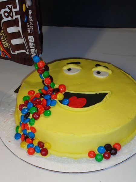 Yellow smiley face cake with M&M's pouring from an open bag and spilling onto the cake.