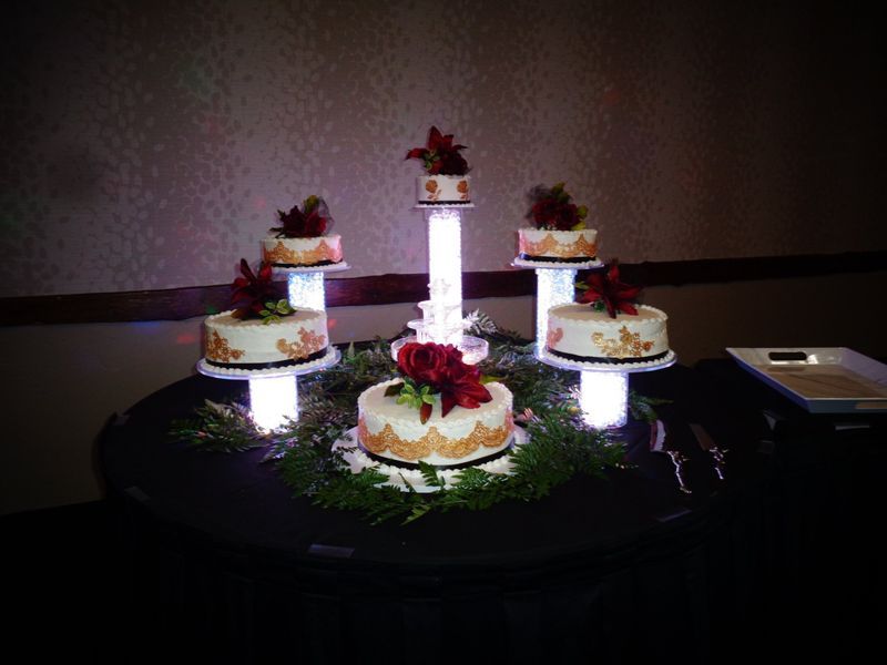 Wedding cake display with six tiers, decorated with flowers and lights. Cakes sit on illuminated pedestals, arranged on a table.