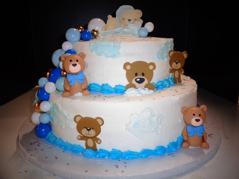Two-tiered baby shower cake with teddy bear decorations, blue and white frosting, and balloon accents.