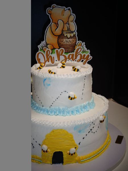 Two-tiered baby shower cake with Winnie the Pooh topper, bees, and beehive. White frosting with blue accents.