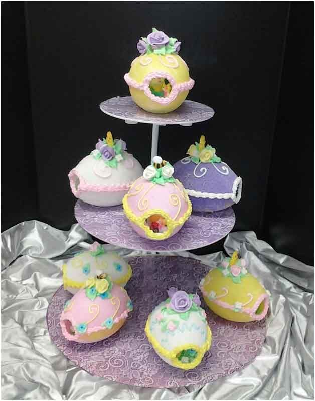 Tiered cake stand with colorful, decorated egg-shaped treats, some with cutouts, topped with flowers.