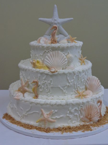 Three-tiered white cake, decorated with seashells, starfish, and seahorses, on a sandy base.
