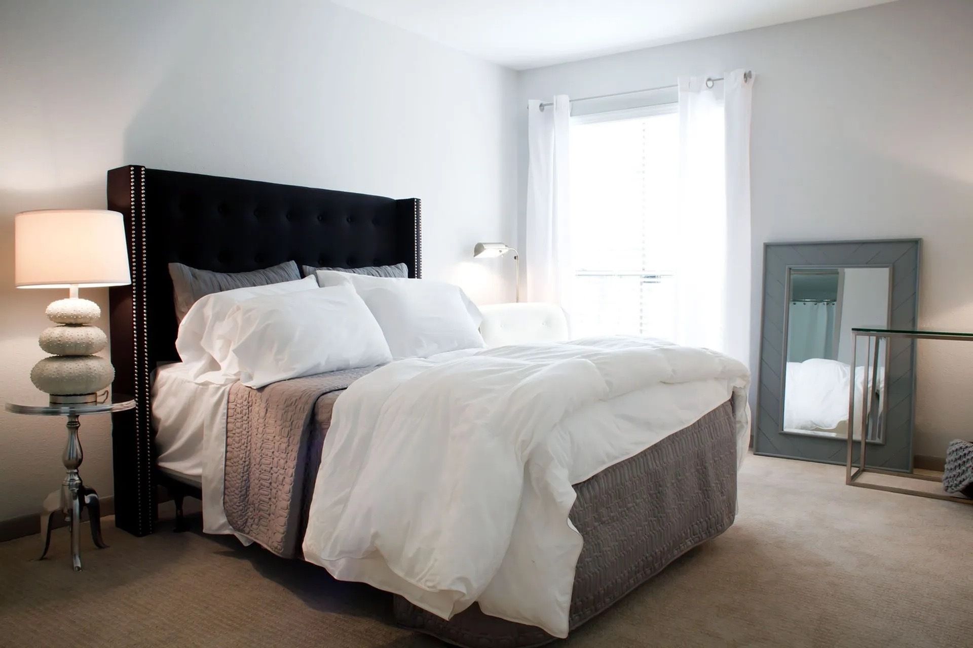 Bedroom with a large tufted headboard, white bedding, window with sheer curtains, and a full-length mirror.
