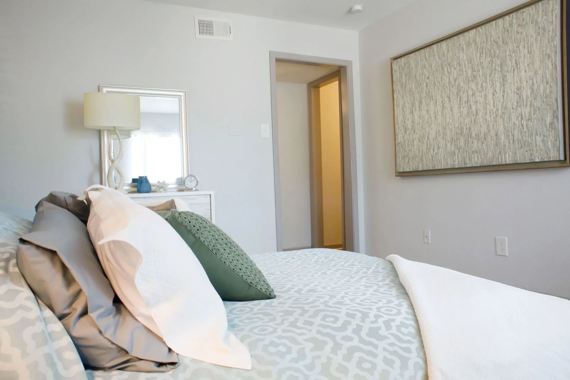 Bedroom in a modern apartment with a bed, dresser, lamp, mirror, and large wall art.