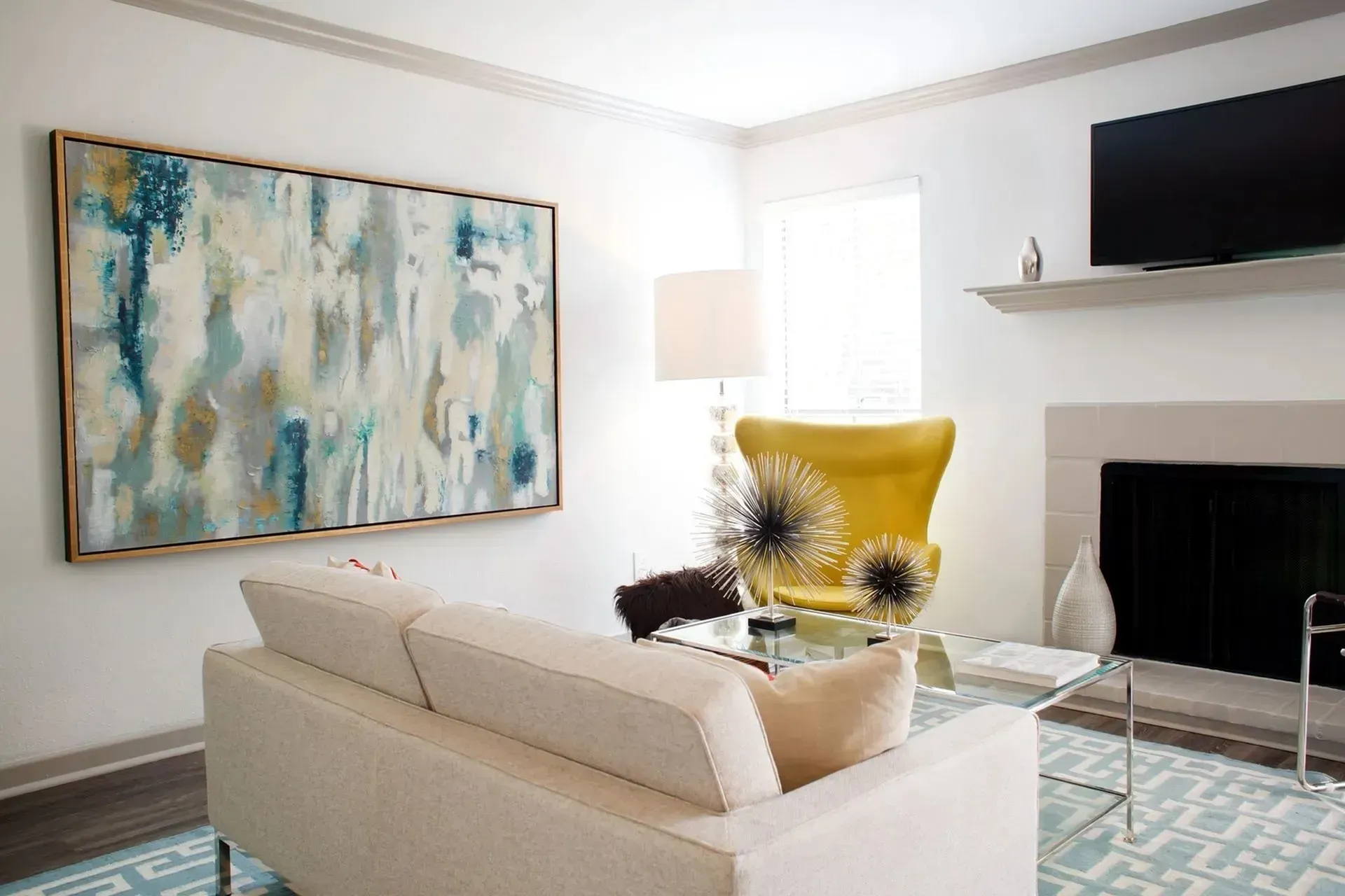 Cozy living room with a beige sofa, glass coffee table, yellow accent chair, and colorful wall art.