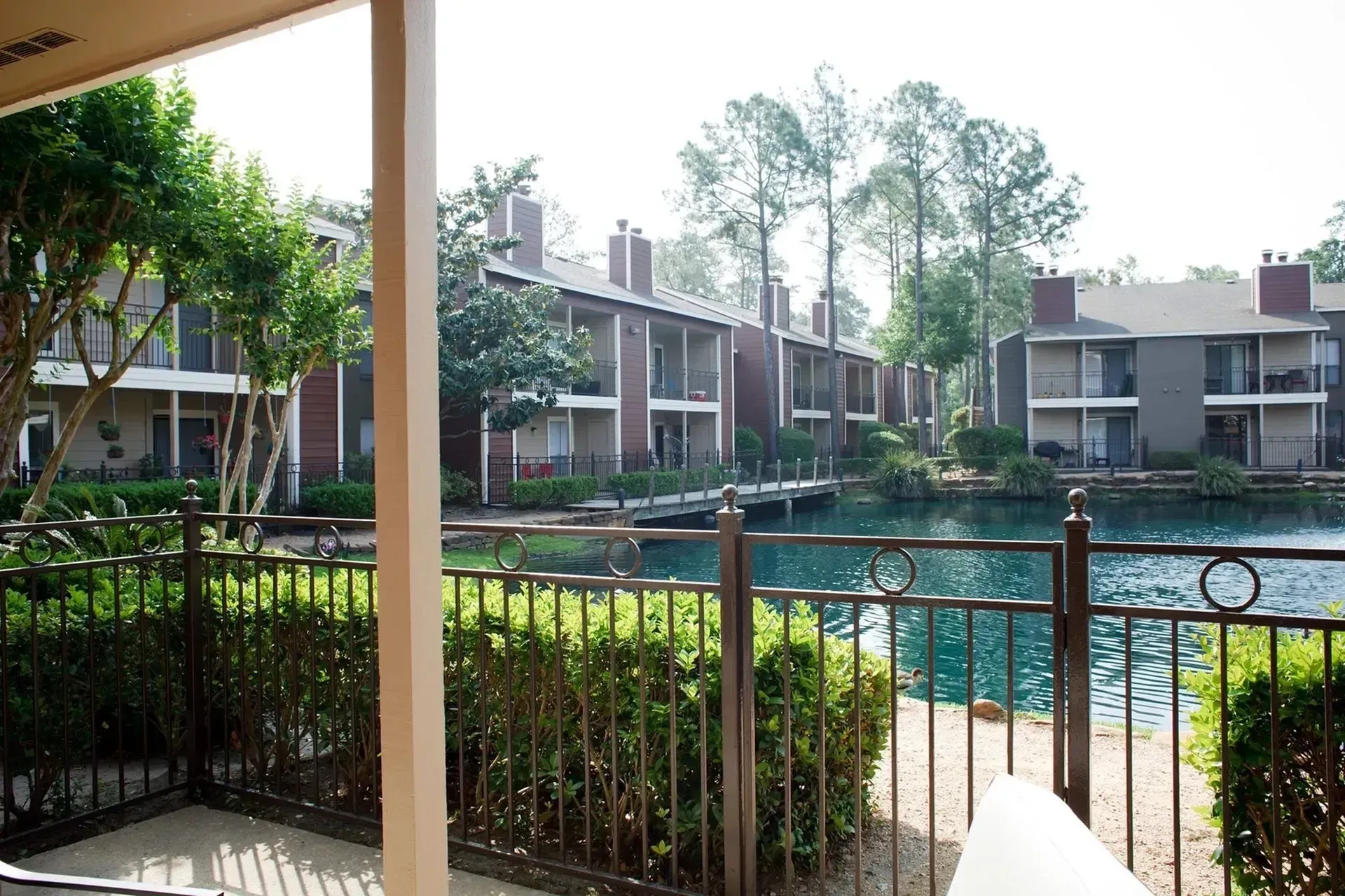 View of apartment buildings by a pond with greenery and a fence in the foreground.