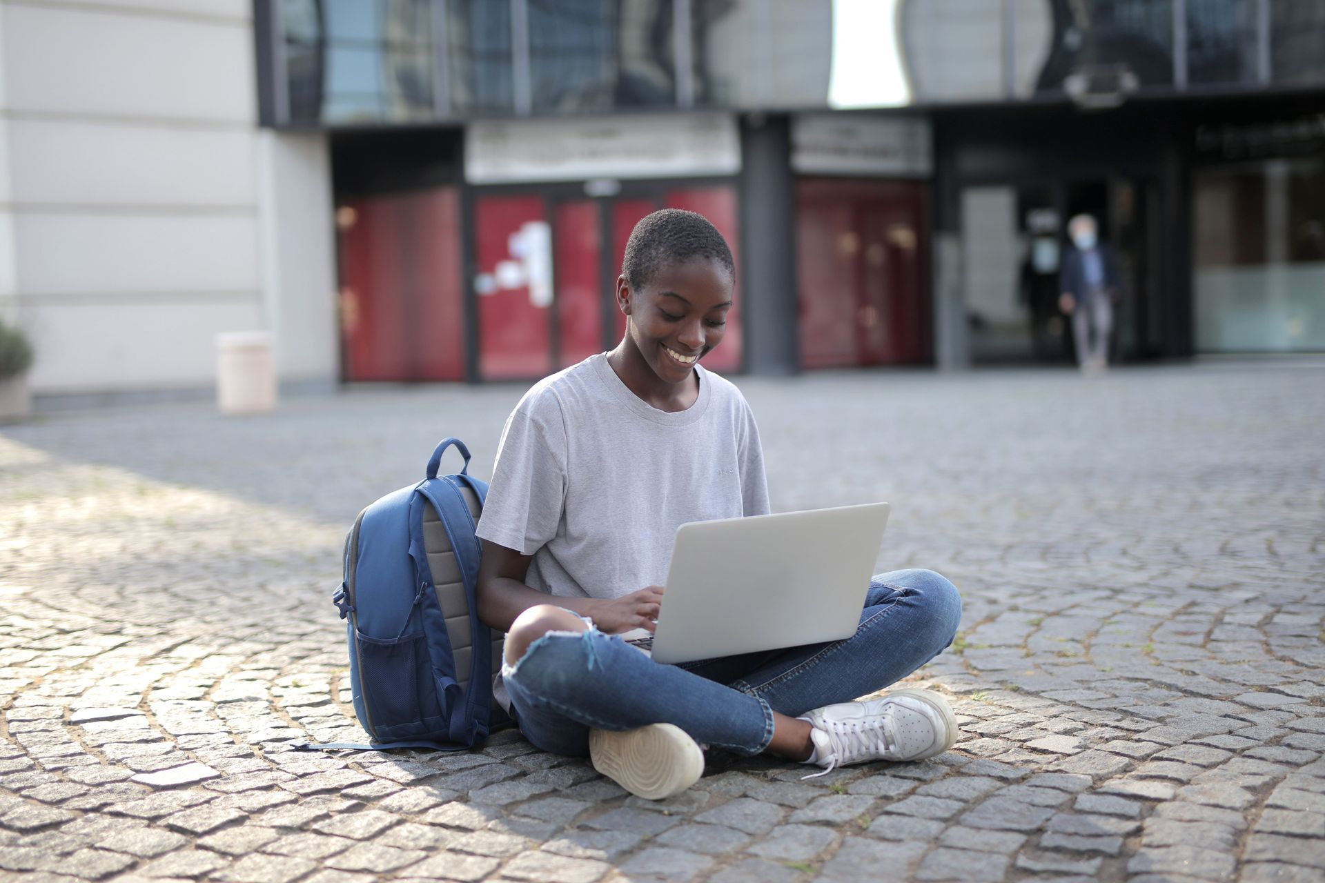 Person sitting cross-legged on a cobblestone plaza using a laptop beside a blue backpack.