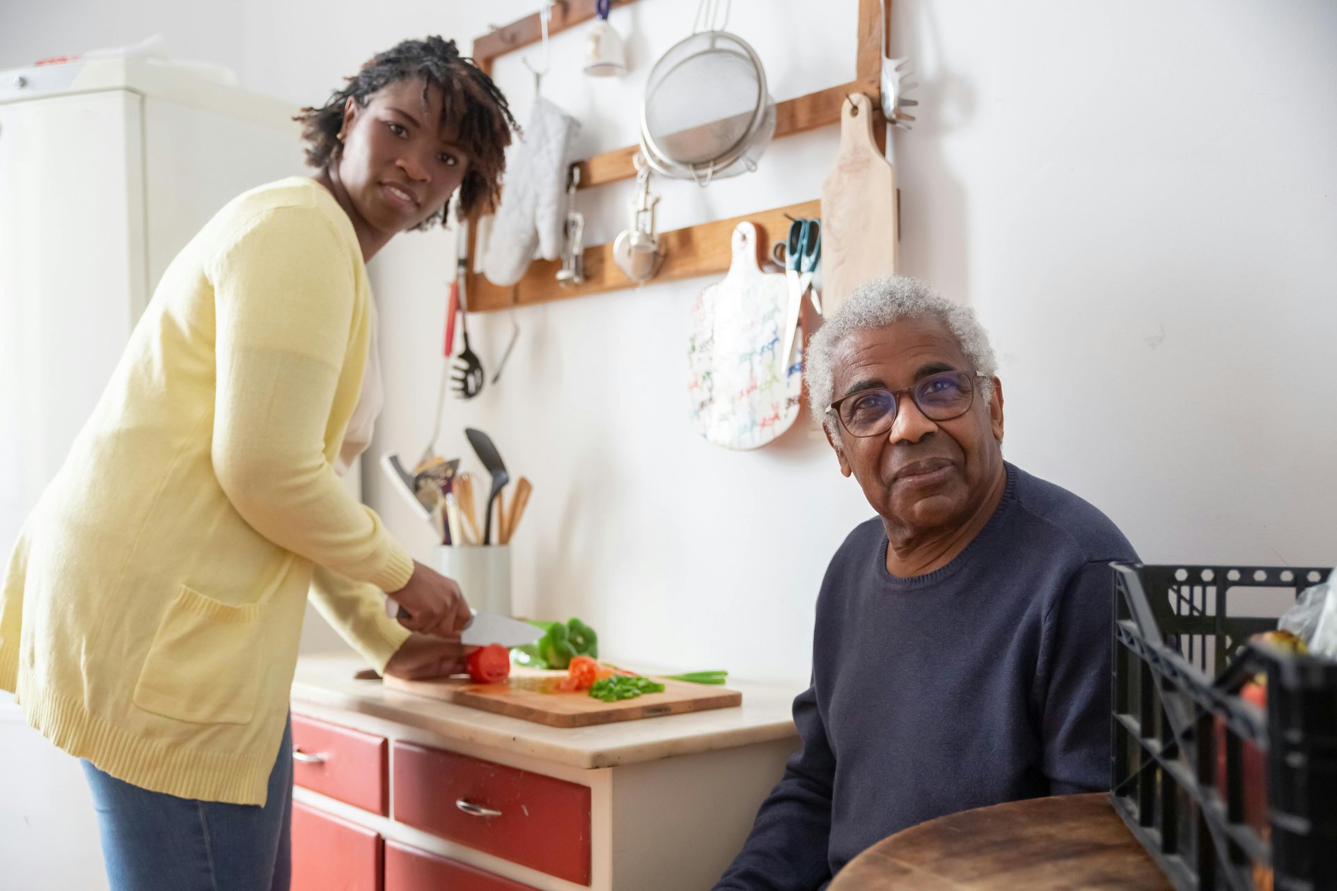 Woman cutting vegetables, older person seated in kitchen.