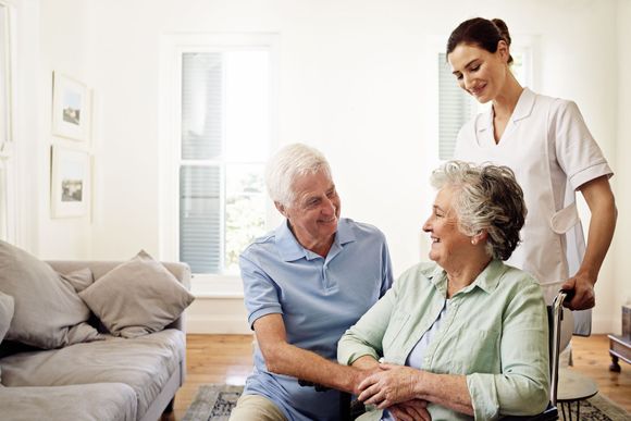 A caretaker assists an elderly woman in a wheelchair, with a man looking on, in a home setting.