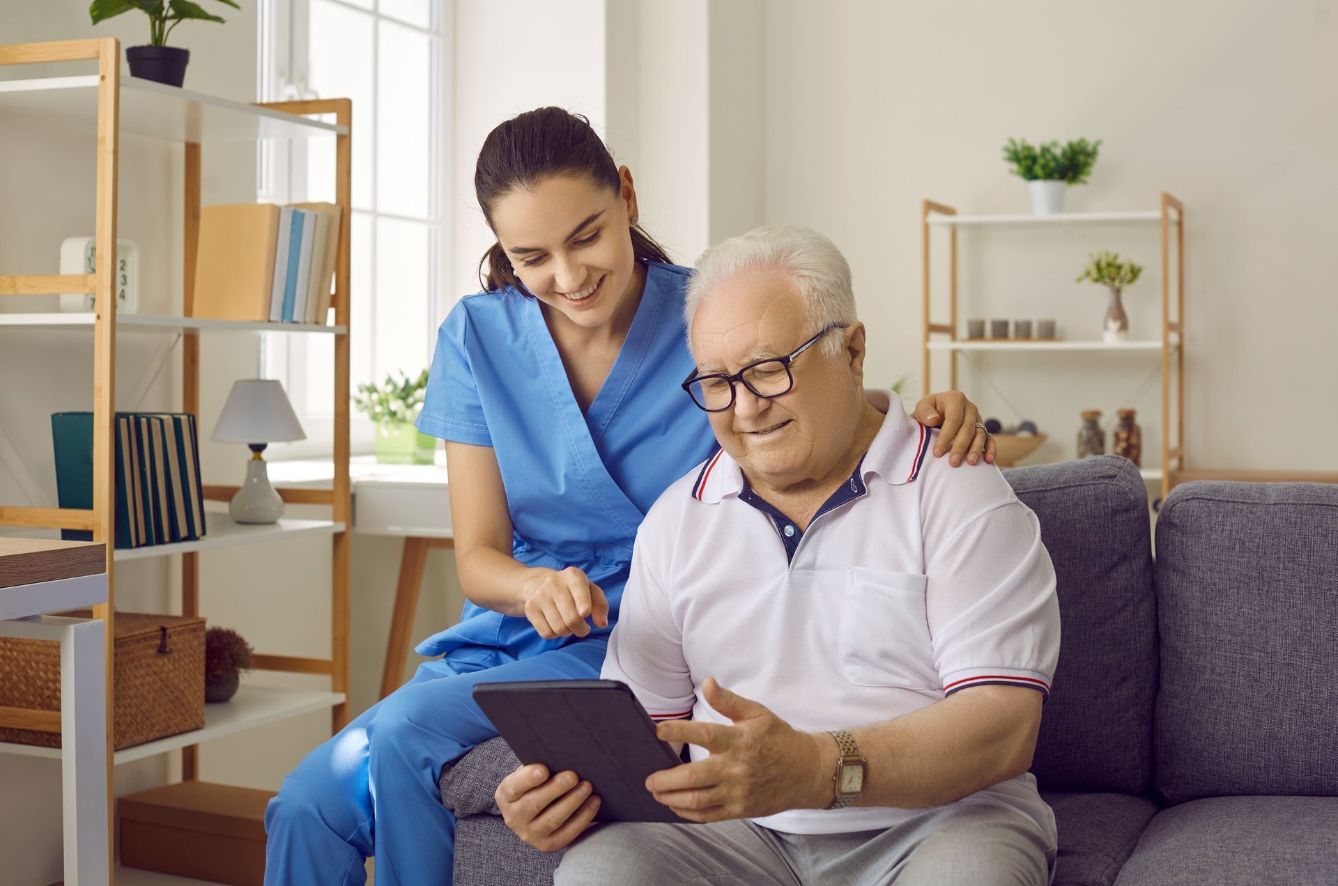 Caregiver assisting an older adult with a tablet on a couch. They are smiling.