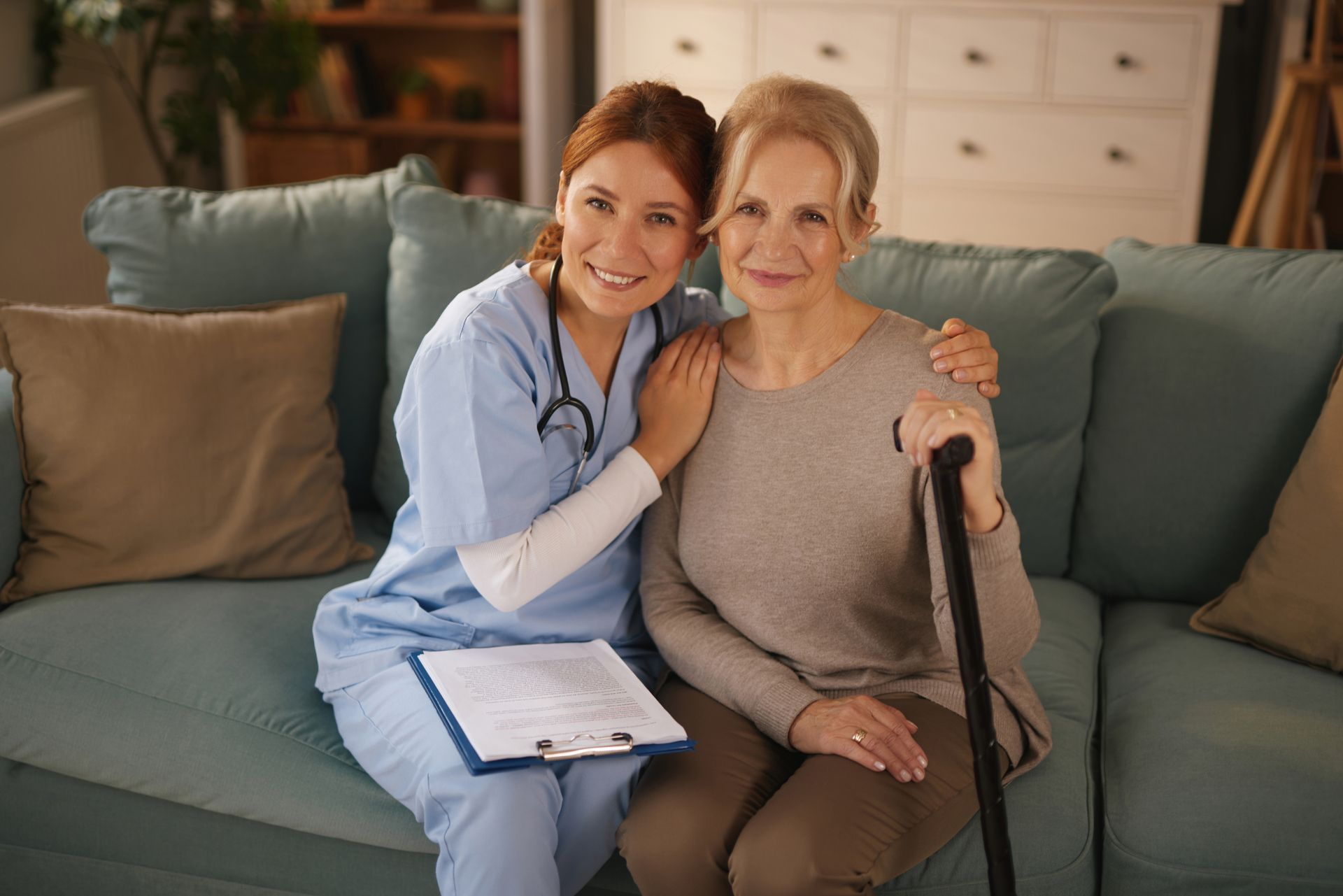 Nurse with a stethoscope, arm around an elderly woman seated on a sofa holding a cane. They are smiling.