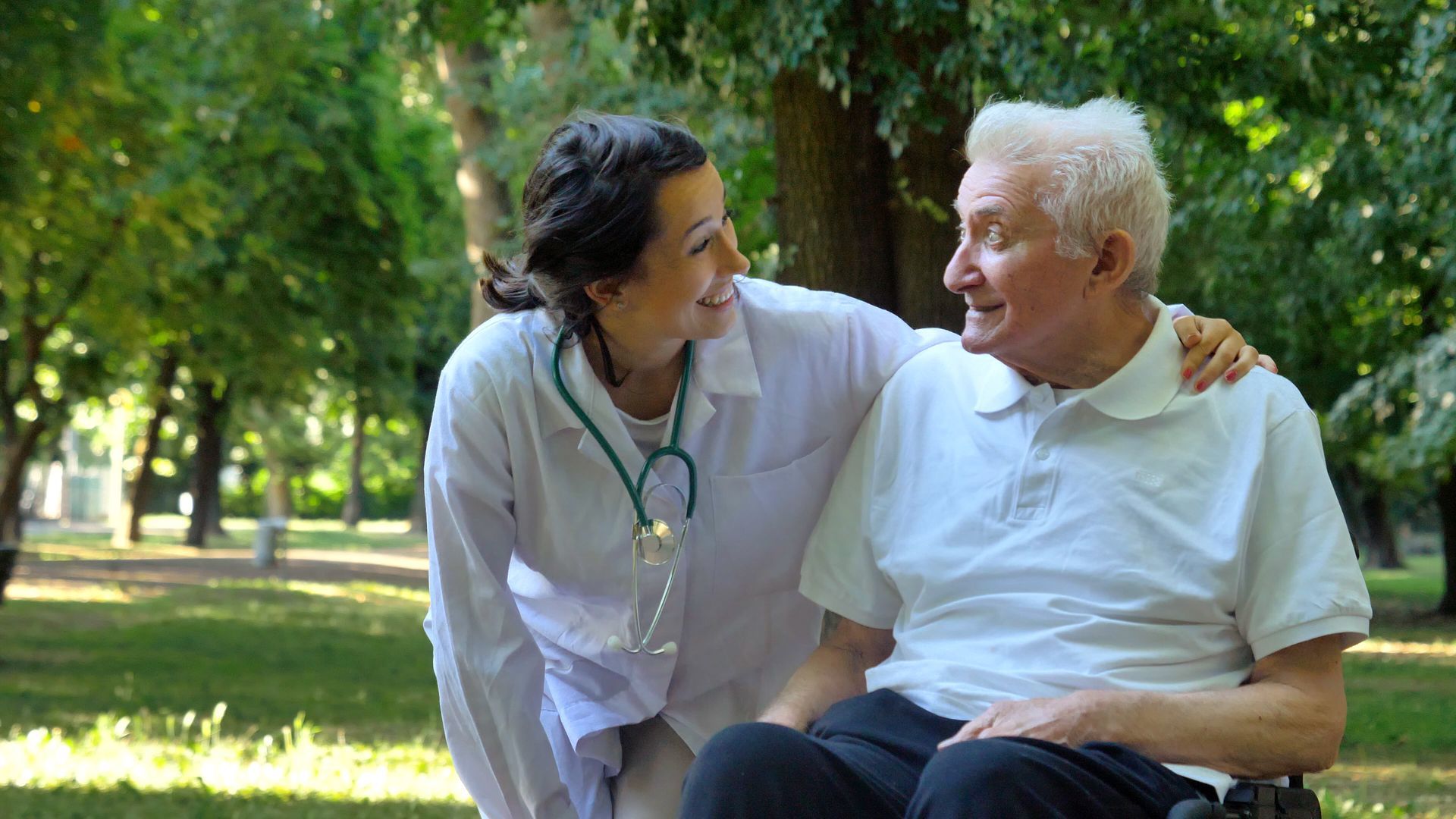 Woman in lab coat with stethoscope, smiles at elderly man in wheelchair outdoors.