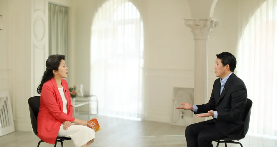 Two people in chairs in a light room. Woman in red jacket, man in suit, talking with hands.