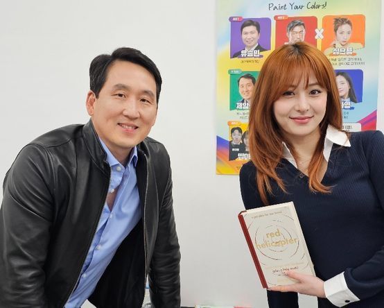 Man in black jacket and woman with red hair holding a book; standing next to a poster; smiling.