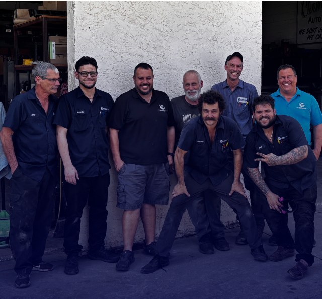 Group of eight men posing together outside a garage. Some are smiling, and one makes a peace sign. | Clemente's Auto Care