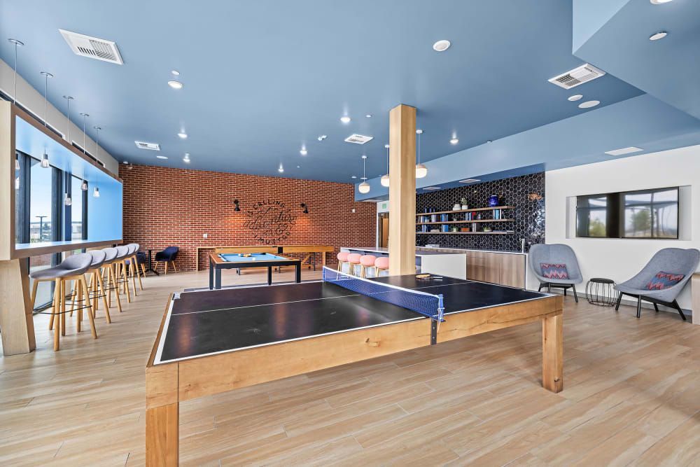 Modern recreation room with a blue ceiling, brick accent wall, and hardwood floors. The space includes bar seating at a high counter, table tennis, and foosball tables, with comfortable lounge chairs and a shelving unit with books and decorative items. Natural light is filtering in through the windows at Marq Iliff Station in Aurora, CO.