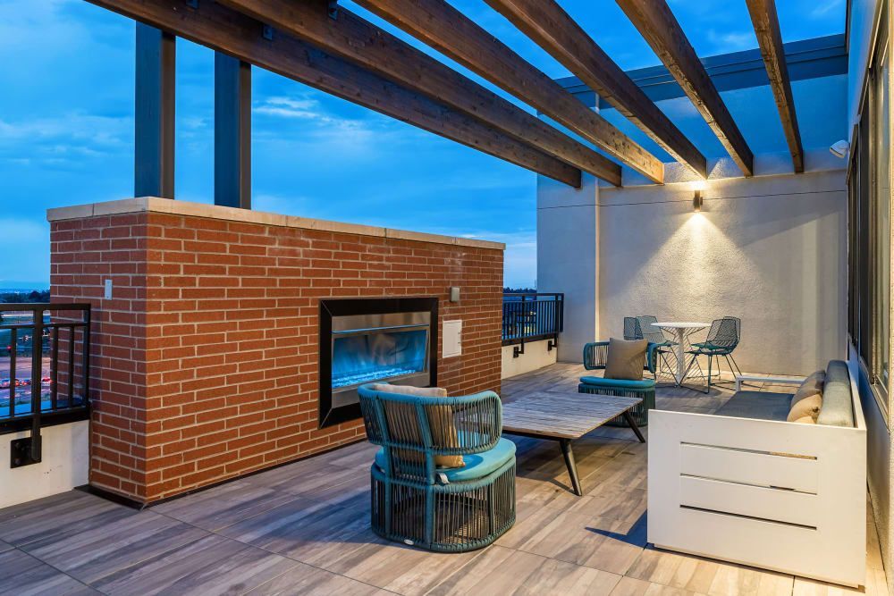 Outdoor residential rooftop patio at dusk with modern furniture, a brick fireplace, wooden pergola structure, and mood lighting. The floor is finished with faux-wood tiles and the space includes a two-seater sofa, two round accent chairs, and a small dining table with chairs. A soft glow is cast from the wall sconce onto the textured white wall. The patio overlooks a view of the surrounding area with visible distant lights at Marq Iliff Station in Aurora, CO.
