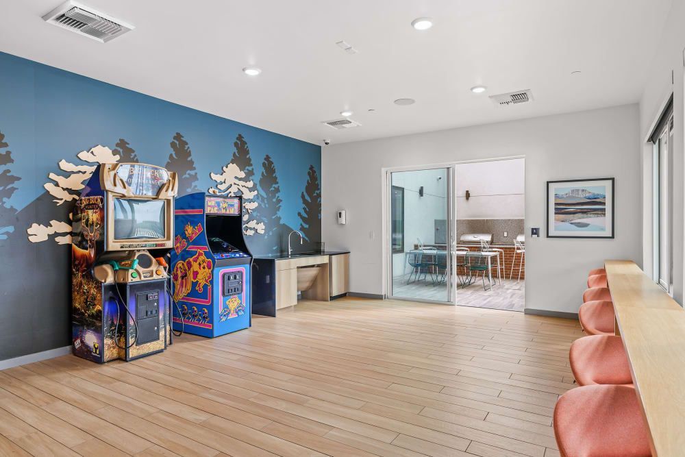 Modern rec area with a recreational corner featuring retro arcade gaming machines against a blue wall with decorative tree silhouettes. The open space leads to a dining area through sliding glass doors, showcasing a contemporary kitchen and a framed scenic artwork on the adjacent wall. Warm wooden flooring extends throughout, complemented by bright, natural lighting at Marq Iliff Station in Aurora, CO.