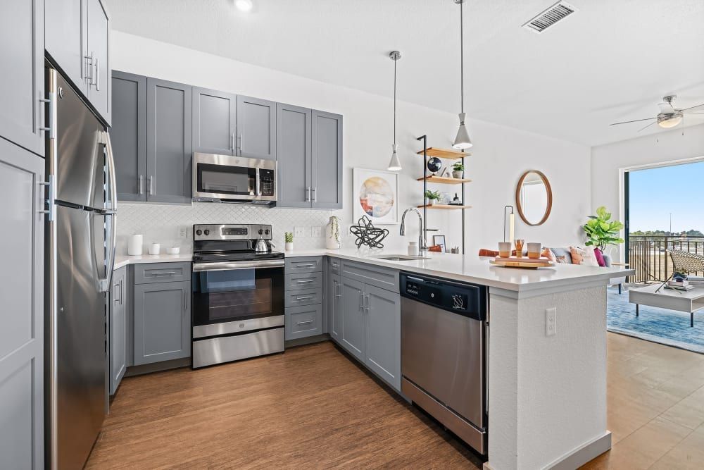 Modern kitchen interior with grey cabinetry, stainless steel appliances, and white subway tile backsplash. Features include a central island with bar stools and pendant lighting above. Hardwood flooring and a balcony with a view are visible in the background at Marq Iliff Station in Aurora, CO.