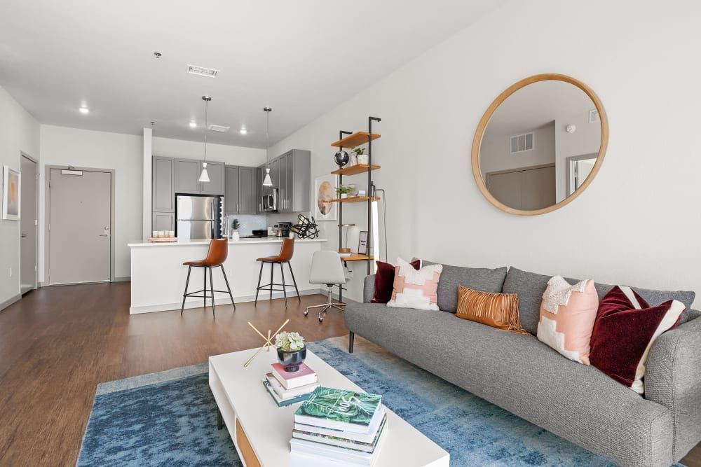 Modern open-concept living room with a gray couch adorned with colorful pillows, a large round mirror on the wall, and an adjoining kitchen with white countertops and bar stools at Marq Iliff Station in Aurora, CO.