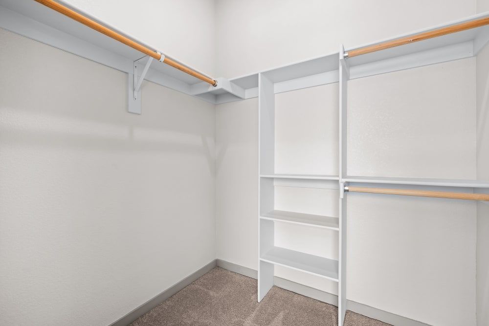 An empty walk-in closet with white walls and beige carpet flooring. Features include built-in white shelves on the right side and two metal closet rods with wooden front bars for hanging clothes at Marq Iliff Station in Aurora, CO.