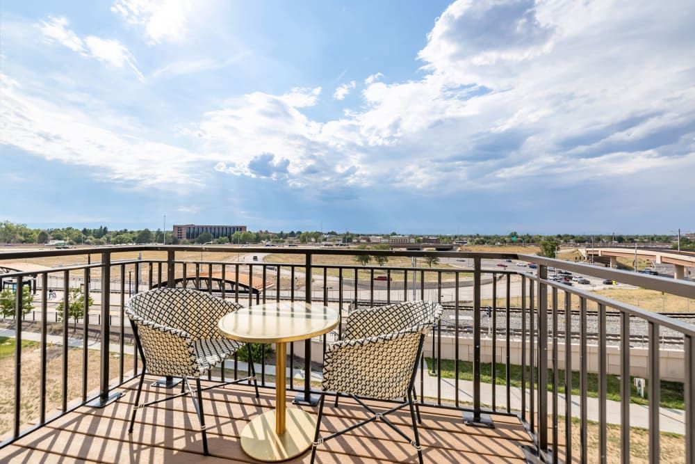Spacious balcony overlooking a highway with a metal railing, featuring a round table and two chairs with patterned cushions under a partly cloudy sky at Marq Iliff Station in Aurora, CO.