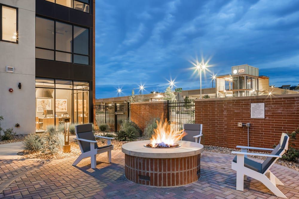Outdoor patio area with a lit fire pit surrounded by three Adirondack chairs in the evening. The space features brick paving and landscaped areas with small bushes and grasses. A glass-fronted building is visible on the left, and there is a brick wall on the right with a sign posted. Starburst effects are created by the bright lights above at Marq Iliff Station in Aurora, CO.