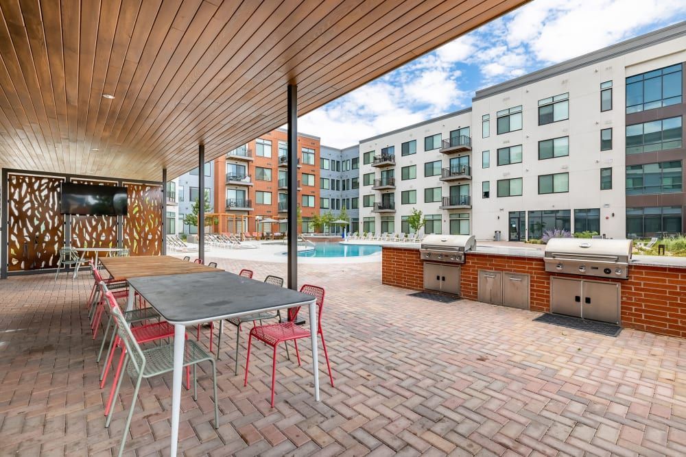 Outdoor residential common area with a covered patio featuring a wooden ceiling, a communal table with red chairs, and an outdoor kitchen with grills. In the background, there's a swimming pool and modern apartment buildings under a cloudy sky at Marq Iliff Station in Aurora, CO.