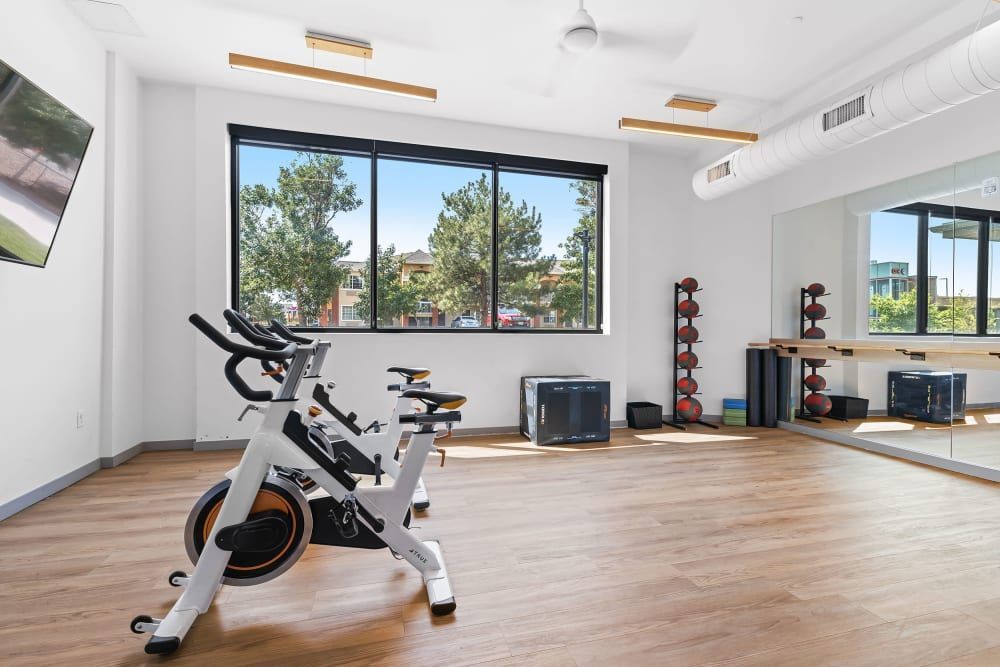 A bright and modern fitness room with large windows, featuring an exercise bike in the foreground, a rack of red and black weights along the wall, and various workout equipment at Marq Iliff Station in Aurora, CO.