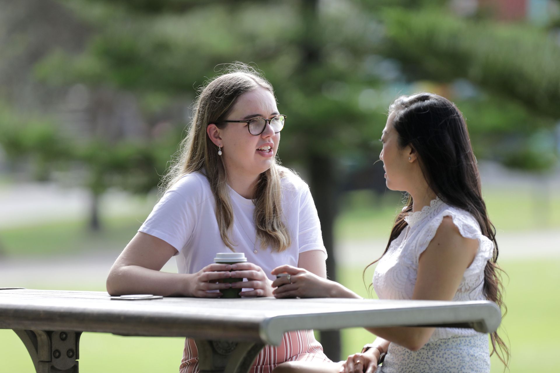 Two young women sitting at a park bench chatting.