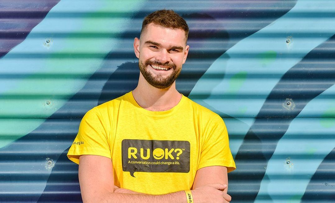 Isaac Humphries, in a yellow R U OK? t-shirt, crosses his arms and smiles