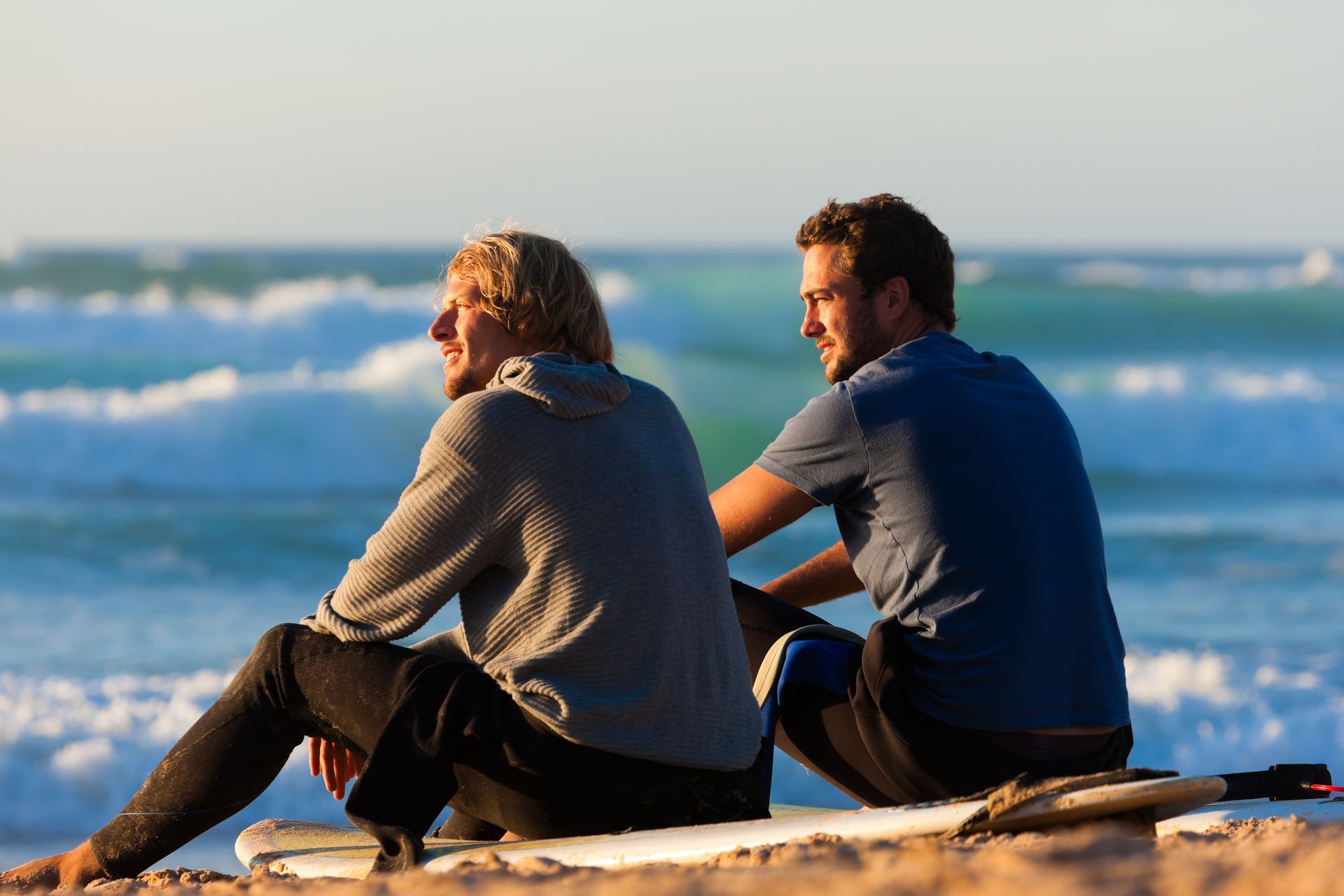 Two young men sit on the sand at a beach and chat.