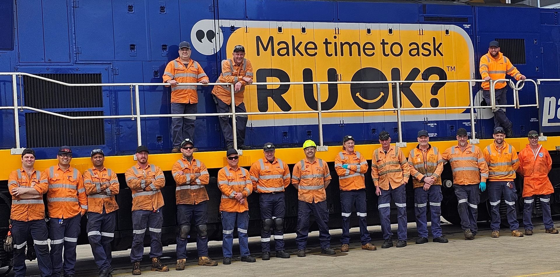 Rail workers stand in front of a train painted with a 'Make time to ask R U OK?' logo.