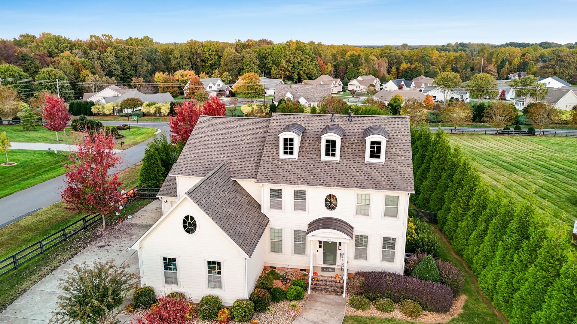 White two-story house with dormers, surrounded by trees with fall colors and a green field.