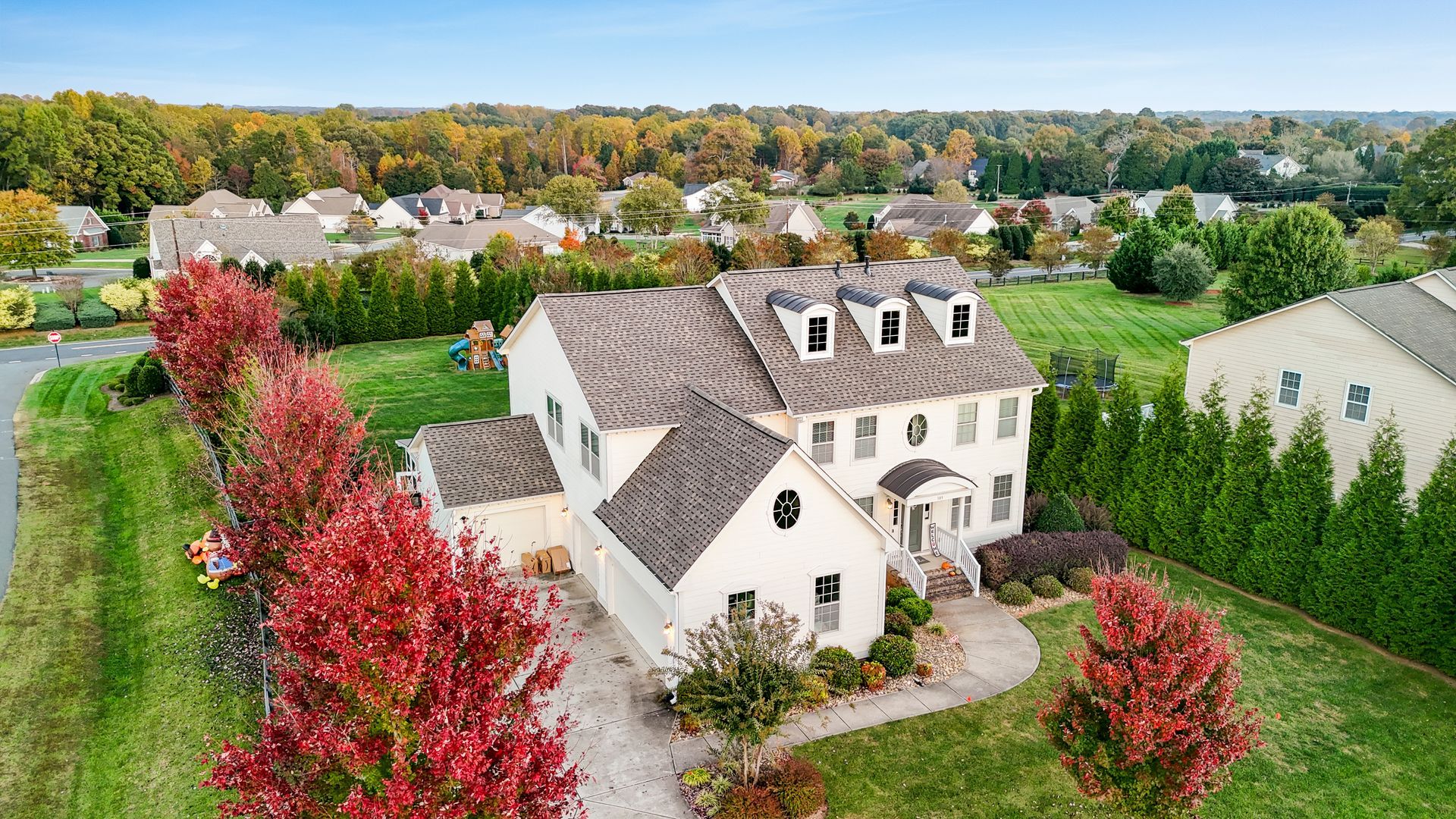 Aerial view of a white two-story house with a brown roof, surrounded by green grass and red trees.