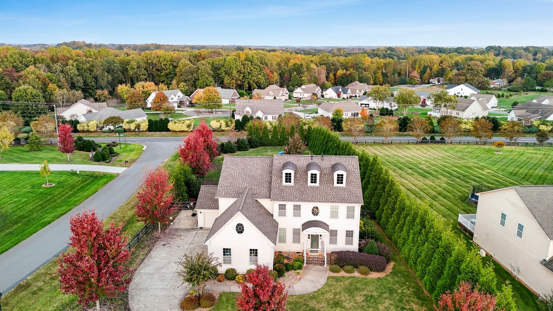 Large, two-story house with a circular driveway, surrounded by greenery and other homes. Autumn colors are visible.