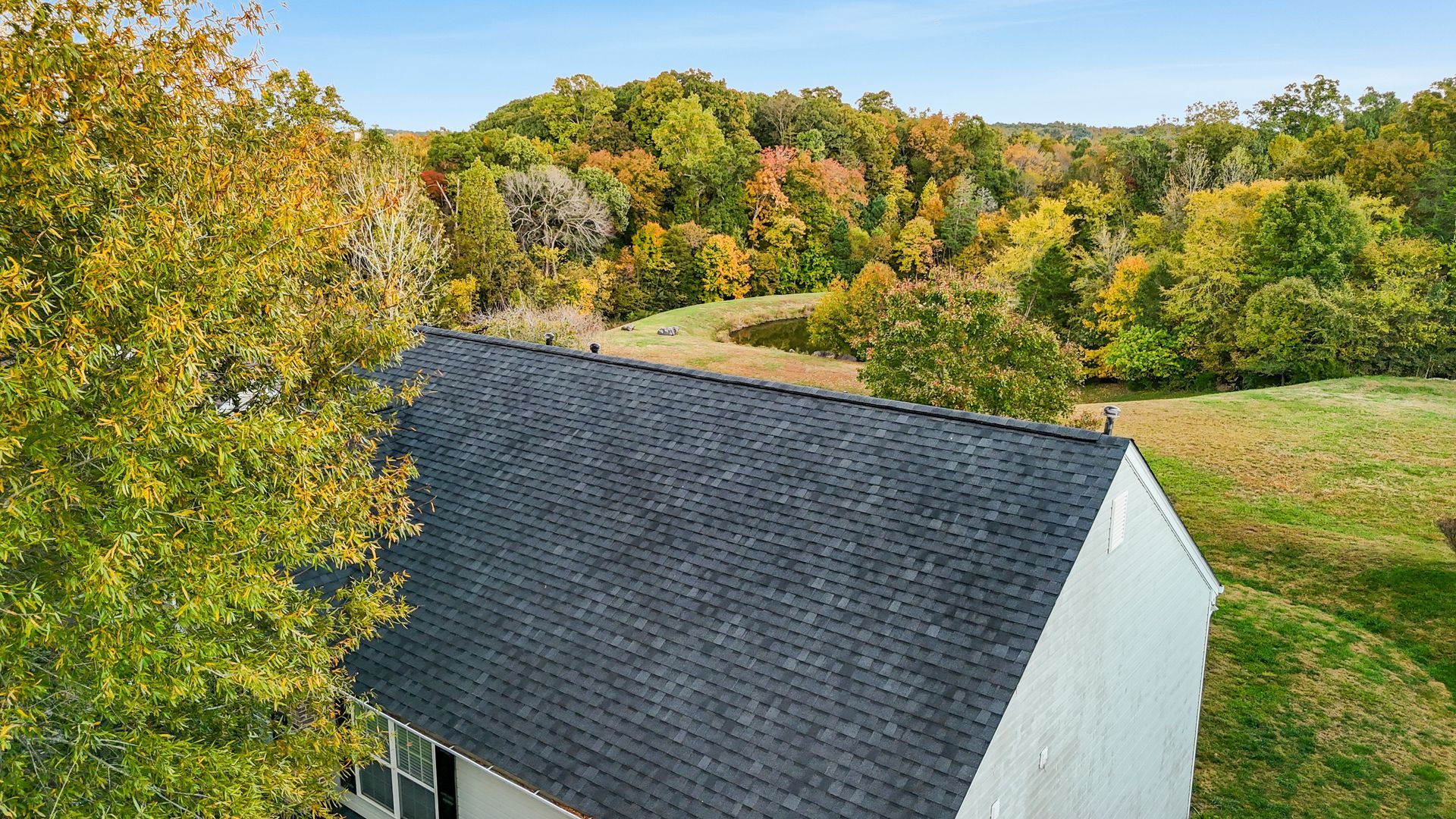 Rooftop of a house with dark shingles, surrounded by trees with autumn foliage.