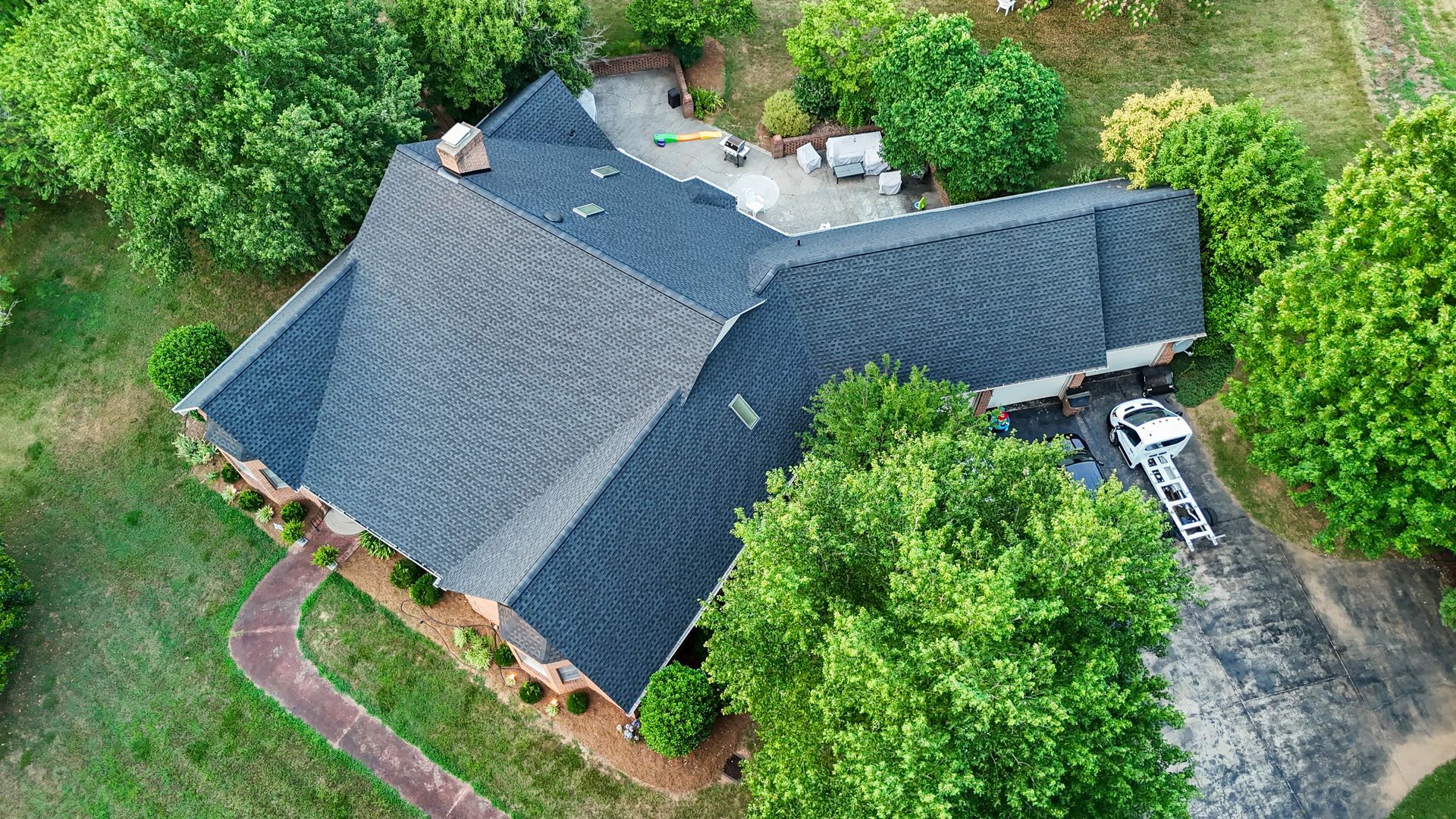 Aerial view of a dark roofed house surrounded by green trees and a driveway with a truck.