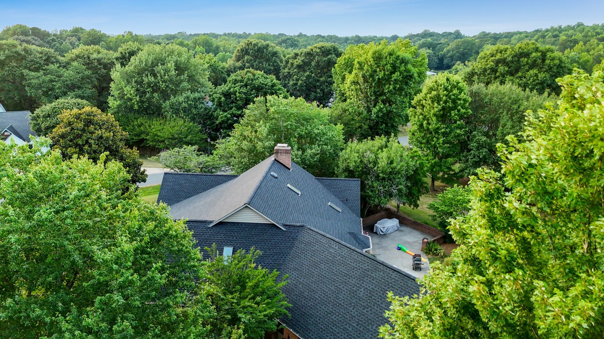Overhead view of a house with a dark roof surrounded by lush green trees, under a blue sky.