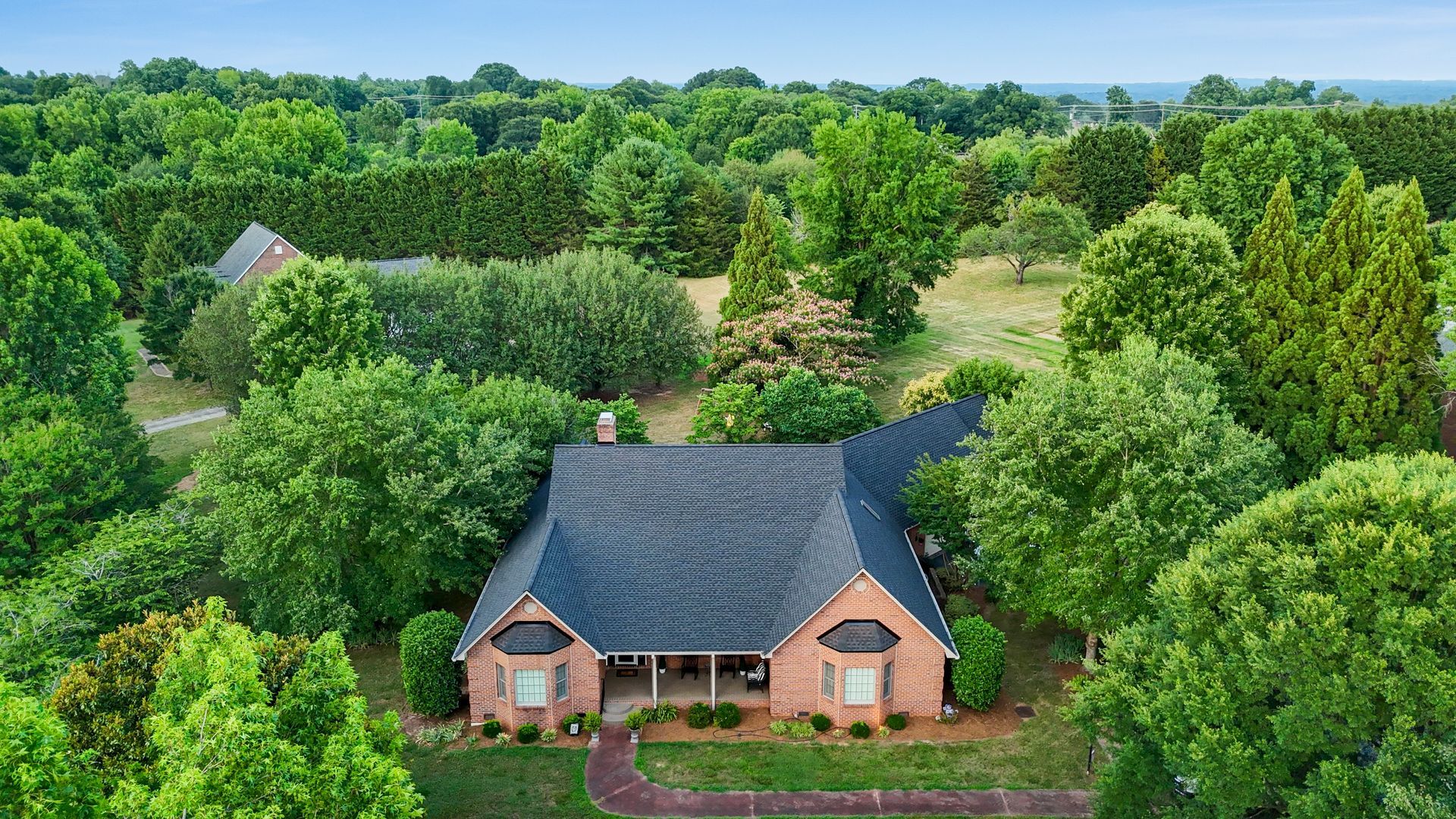 Brick house with dark roof surrounded by lush green trees, in an outdoor setting.