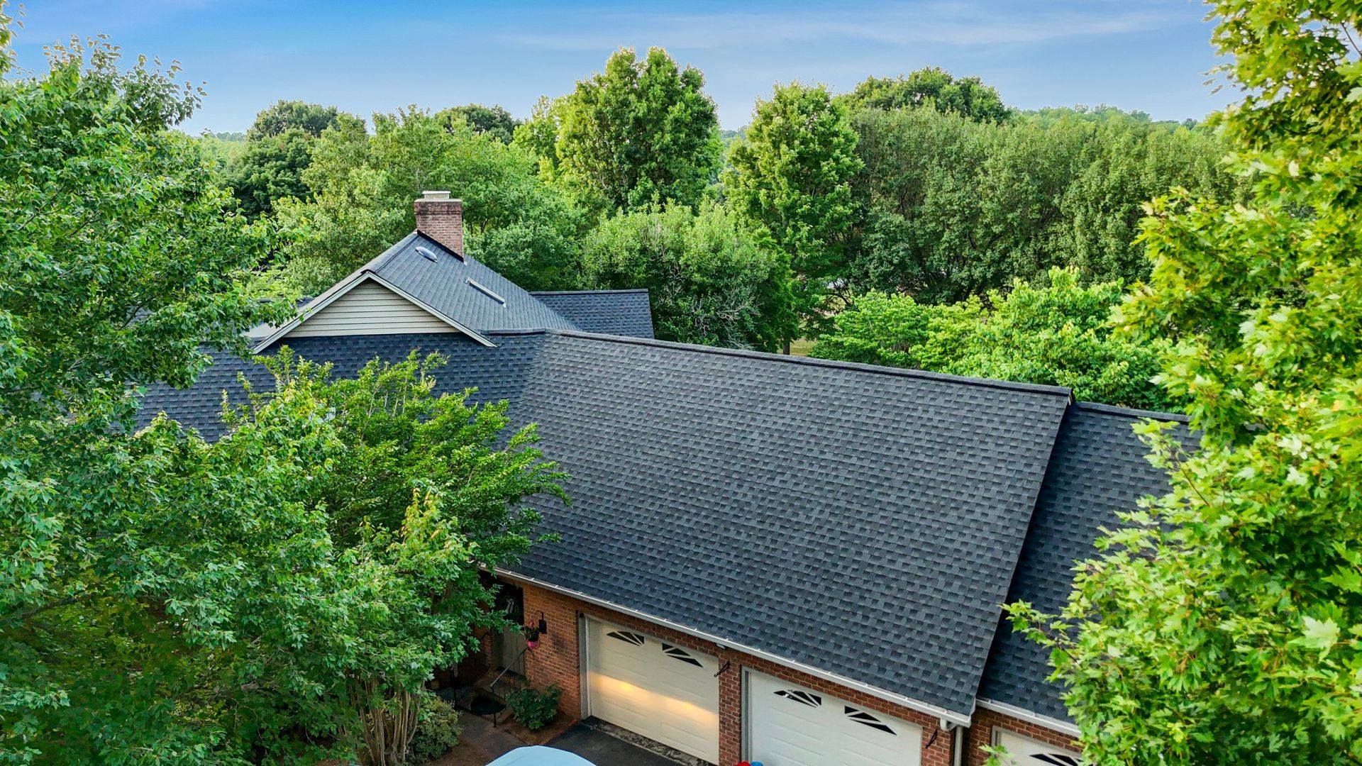 Aerial view of a house with a dark gray roof surrounded by lush green trees. Three garage doors are visible.