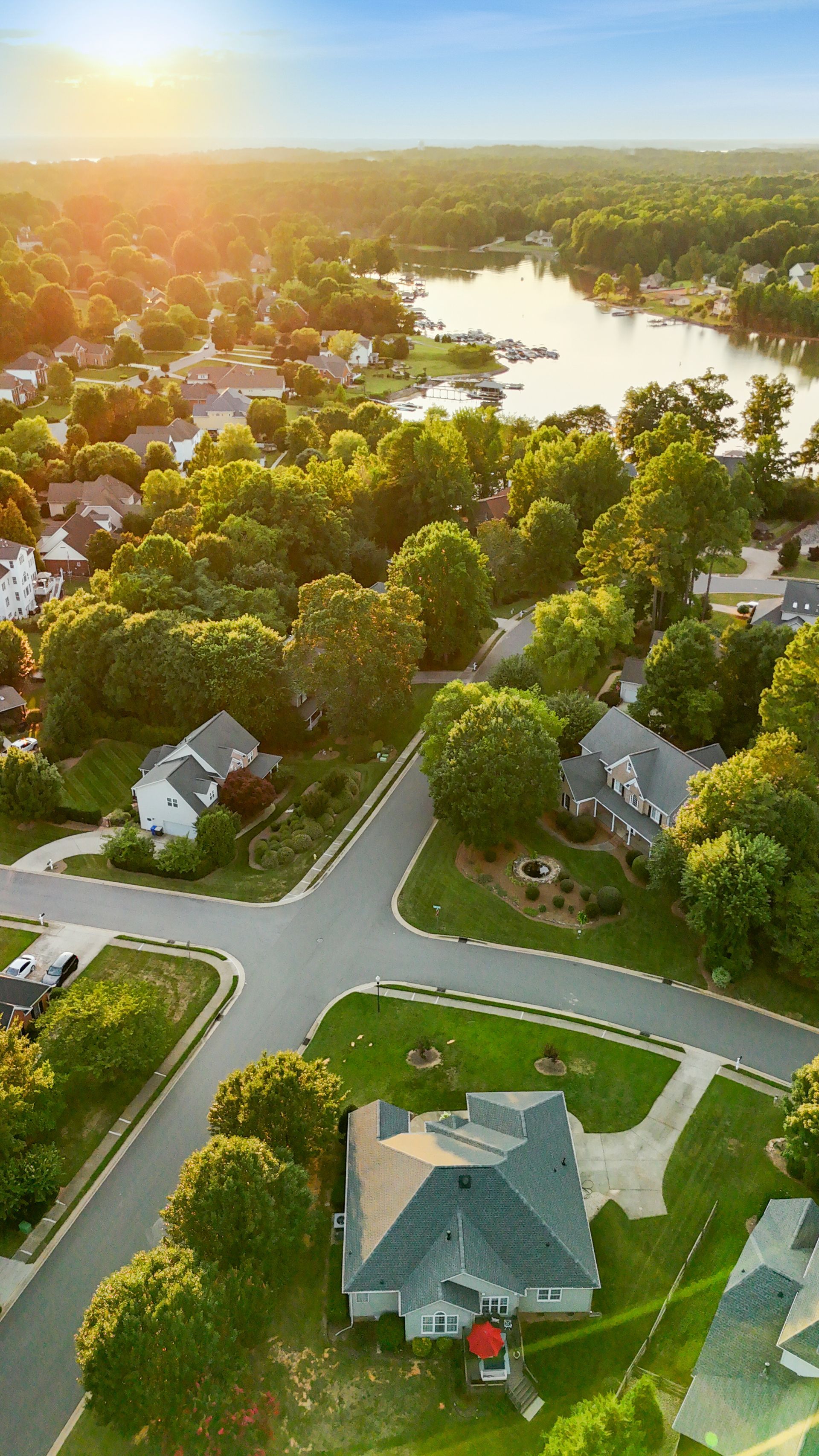 Aerial view of a suburban neighborhood with houses, streets, trees, and a lake, lit by the setting sun.