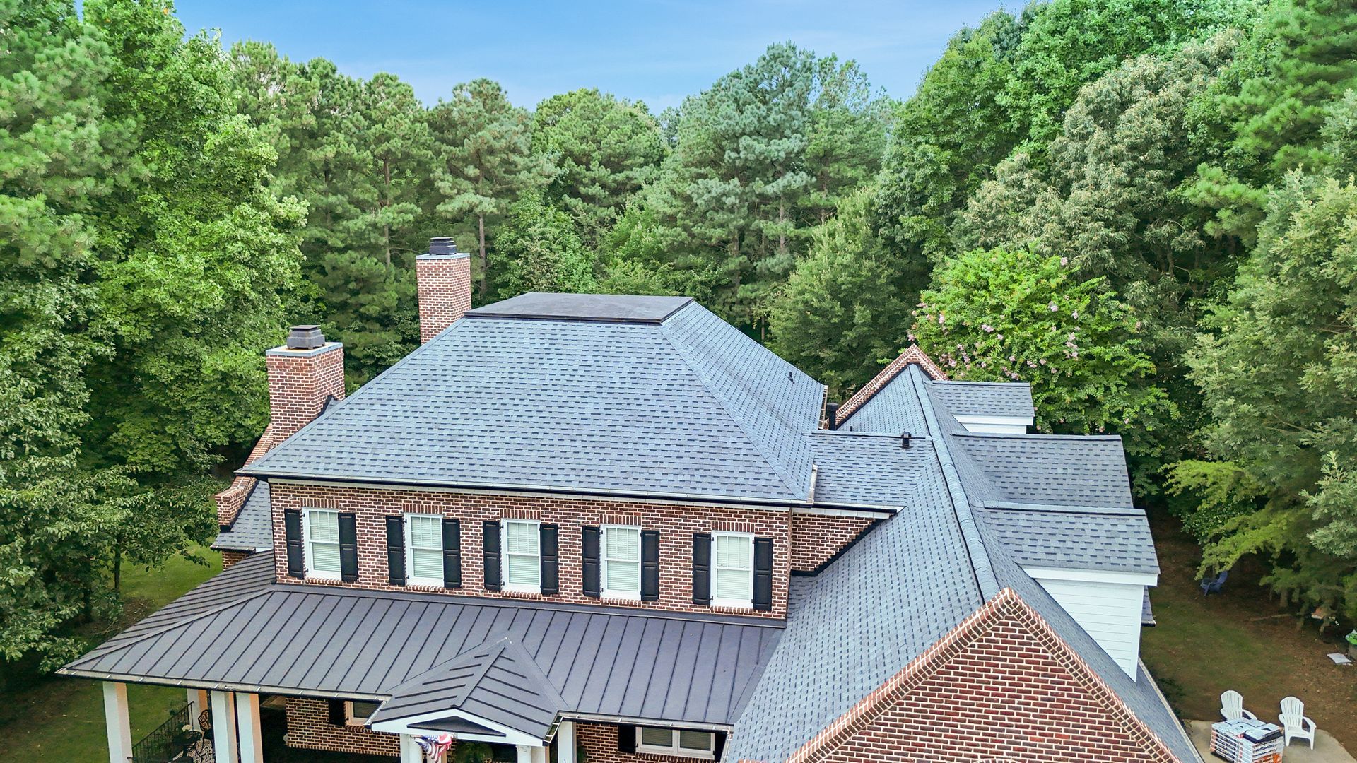 Brick house with dark roof and multiple dormers, surrounded by green trees.